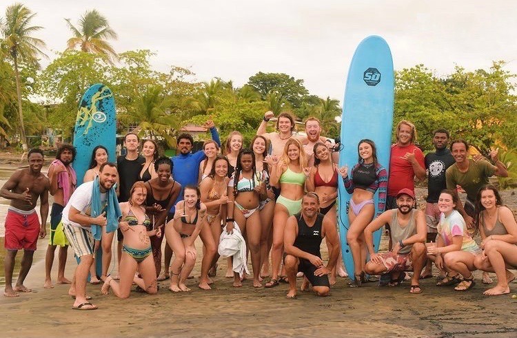 A group of smiley students pose on the beach with surf boards
